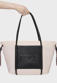 Czarna torba typu tote w jasnoróżowym kolorze z teksturowanym skórzanym przodem, z wytłoczonym logo "TOUS SINCE 1920" oraz dwoma długimi uchwytami. Pleciony detal akcentujący.