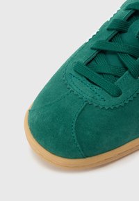 Πράσινο σουέντ �αθλητικό παπούτσι με σόλα από καουτσούκ χρώματος μαστίχας, με επίπεδα κορδόνια και ραφή κατά μήκος του πάνω μέρους.