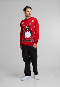 Piros pulóver, amelyen egy napszemüveget, masnival és karácsonyi sapkával ellátott pingvin grafika látható, fehér hópelyhekkel díszítve, valamint a "MERRY X-MAS" felirattal.