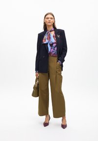 Blazer nero, blouse floreale nei toni di blu e viola, pantaloni larghi oliva, cintura bordeaux, borsa verde, tacchi viola.