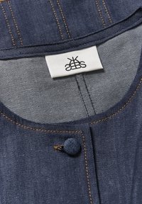 Colletto di capo in denim con chiusura a bottone rotondo, cuciture gialle e un'etichetta con marchio. Il tessuto è di colore blu scuro con una consistenza liscia.