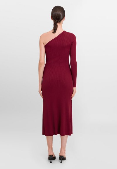 Robe bordeaux côtelée avec un design à une épaule, une manche longue d'un côté et une longueur mi-mollet, agrémentée de chaussures à talons.
