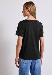 Černé tričko s krátkým rukávem a širokým výstřihem, vyrobené z měkké látky, nošeno s světle modrými džíny. Na zápěstí je viditelný zlatý náramek.