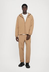 Bež jakna na patentni zatvarač s kapuljačom preko bijele majice, uparena s odgovarajućim bež cargo hlačama. Crne cipele zaokružuju odjevnu kombinaciju.