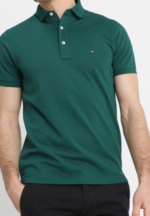 Mann trägt ein dunkelgrünes kurzärmeliges Polohemd mit weißen Knöpfen und kleinem Logo auf der linken Brust, kombiniert mit schwarzen Hosen.