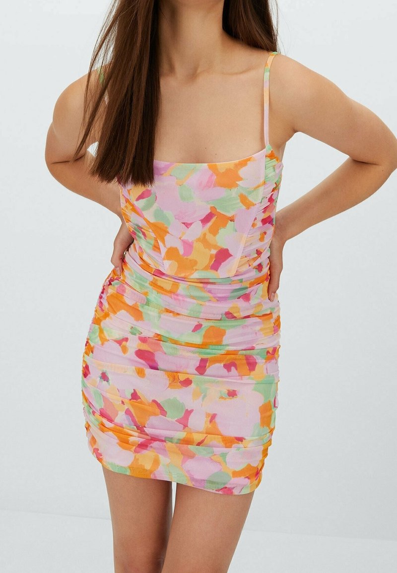 Femme portant une mini-robe sans manches froncée, avec de fines bretelles et un motif floral abstrait multicolore vif.
