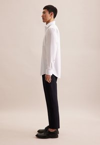 Chemise blanche à manches longues avec une poche poitrine, assortie à un pantalon noir ajusté et des chaussures en cuir noires, vue de côté.