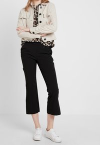 Beżowa krótka kurtka z dwoma przednimi kieszeniami, zestawiona z czarną bluzką w panterkę i czarnymi spodniami w fasonie 3/4, uzupełniona białymi sneakersami.