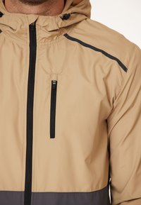 Helle beige Kapuzenjacke mit einem schwarzen Reißverschluss, ausgestattet mit einer Reißverschlusstasche auf der Brust, einem grauen Kontrast im unteren Abschnitt und schwarzen Akzentstreifen.