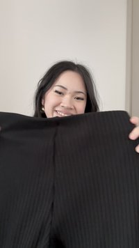 Mladá žena sa usmieva a drží čierne rebrované nohavice na jednoduchom svetlom pozadí.