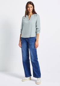 Blouse verte claire à manches longues avec un col en V et des épaules froncées, associée à un jean en denim bleu ample et des baskets blanches sur un fond uni.