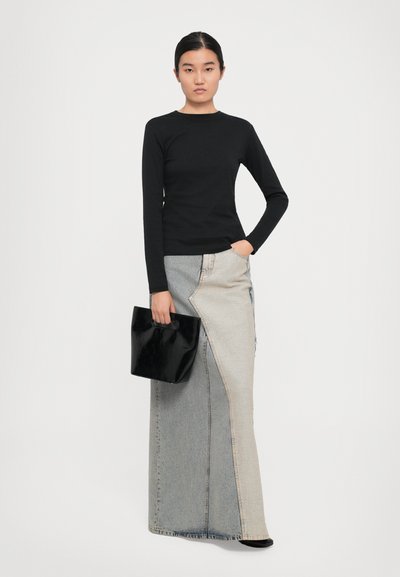 Kvinna i svart långärmad topp och tvåtonad denim maxi kjol, som håller en svart handväska, stående mot en vit bakgrund.