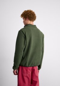 Fleece jacket in dark green with a high collar, textured fabric, and relaxed fit, paired with loose-fitting pink pants.