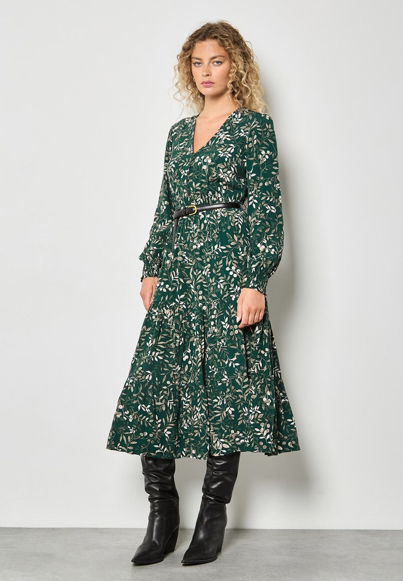 Groene jurk met lange mouwen en een bloemenpatroon, V-hals, gelaagde rok en een getailleerde taille met een zwarte riem. Gecombineerd met kniehoog zwarte laarzen.