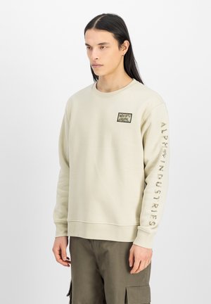 Creme sweatshirt med lange ærmer, prydet med et firkantet logo-patch på brystet og "ALPHA INDUSTRIES" trykt ned ad venstre ærme. Blød tekstur.