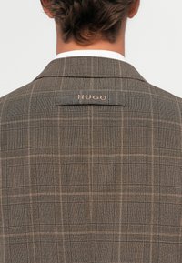 Mann in braunem kariertem Jacket mit "HUGO"-Label auf dem oberen Rücken, stehend vor einem schlichten weißen Hintergrund.