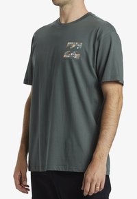 Camiseta de color verde oscuro hecha de algodón, con un diseño gráfico multicolor en la parte frontal. Mangas cortas y cuello redondo estándar.