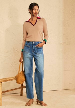 Frau trägt einen beigen Pullover mit buntem Kragen und Bündchen, hoch geschnittene blaue Jeans, hält eine beigefarbene geflochtene Handtasche und steht an einer Holzbank.