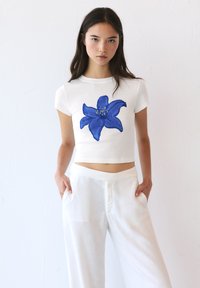 T-shirt bianco accorciato con un design di fiore blu al centro, realizzato in materiale morbido. Abbinato a pantaloni bianchi leggeri.