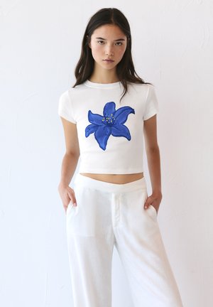 White cropped t-shirt featuring a blue flower design at the center, made from soft material. Paired with lightweight white trousers.