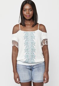Top bianco scollato sulle spalle con ricami floreali blu, dettagli in pizzo e frange di perline sulle maniche. Indossato con pantaloni shorts in denim chiaro.