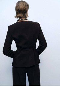 Sacou negru, croi pe corp, cu talie peplum, mâneci lungi și textură fină, asortat cu pantaloni evazați, văzut din spate.