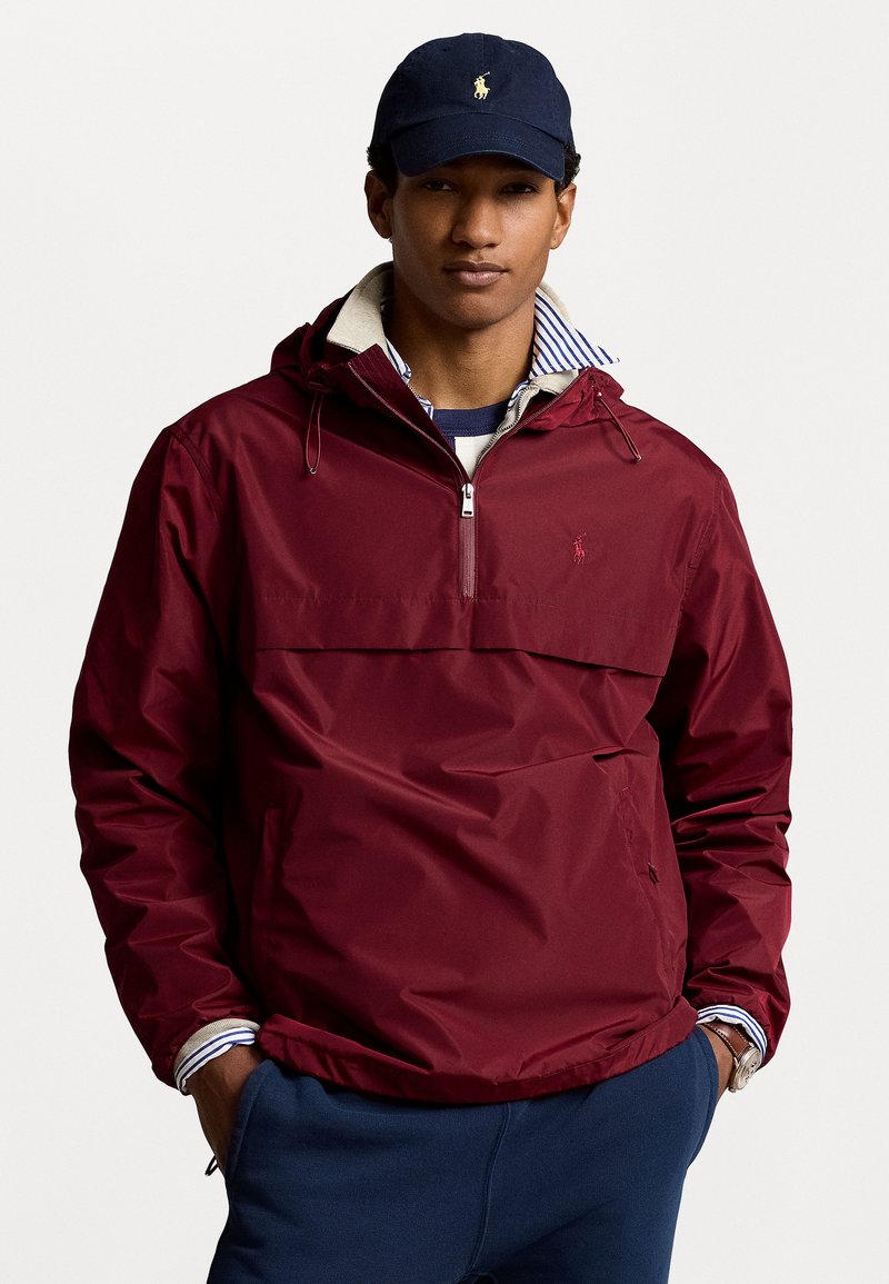 Junger Mann in einem burgunderfarbenen Kapuzenpullover mit einem Viertelreißverschluss, einem gestreiften Kragenhemd, einer marineblauen Cap und marineblauen Hosen mit Händen in den Taschen.