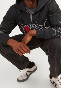 Sudadera con cremallera negra y diseño de telaraña blanca con acentos rojos, fabricada en un tejido suave. Combinada con pantalones negros y zapatillas gris.