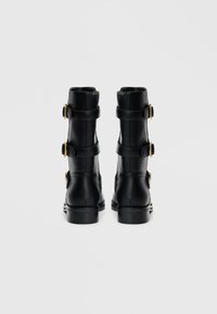 Botas de piel negras hasta el tobillo con dos correas ajustables y hebillas en tono dorado, tacón cilíndrico y textura suave. Se muestra la vista trasera.