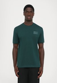Tumši zaļa īsroku T-krekls no mīksta materiāla, ar taisnstūra formas uzšuvumu un logotipa detaļu kreisajā krūšu daļā.