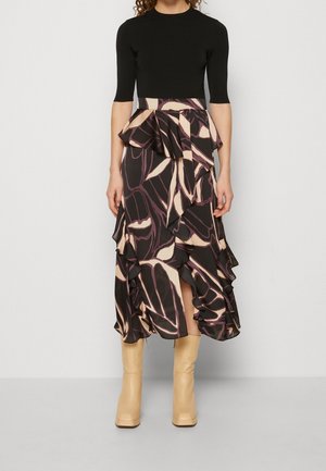 Femme portant un haut noir ajusté et une jupe midi à volants à taille haute avec des motifs floraux beige et violet foncé, assortie à des bottes beiges.