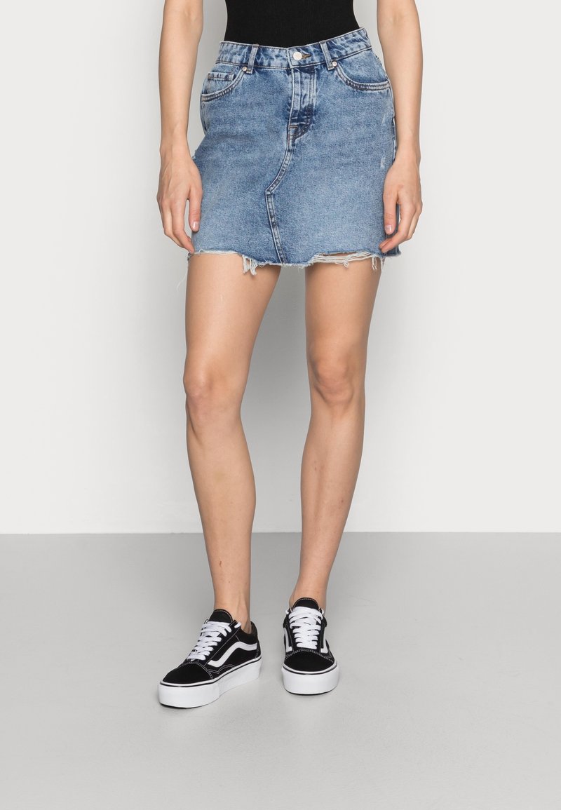 Femme portant une mini-jupe en jean bleu avec un ourlet effiloché, un haut noir et des baskets noires et blanches, debout devant un fond uni.