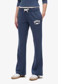 Marineblå sweatpants lavet af blødt stof, med snor i livet og hvidt trykt tekst "SUPER 1985" på venstre lår.