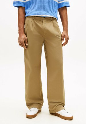 Homme portant un pantalon large couleur sable, des baskets blanches avec des semelles en gomme, et un pull tricoté à rayures bleues sur un fond blanc.