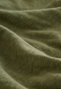 Tykt, blødt stof i en dæmpet olivengrøn farve, med en struktureret vævning og synlige folder, der viser variationer i lys og skygge.