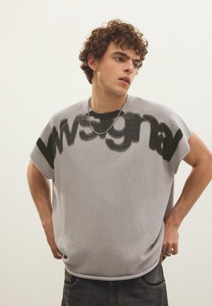 Fiatal férfi göndör hajjal, aki szürke kötött ujjatlan felsőt visel nagy fekete felirattal, fekete farmert, ezüst láncos nyakláncot és gyűrűket.