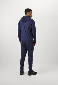 Marinblå sportoverall med huva; har blå detaljer, långa ärmar och avsmalnande byxor. Tyget ser mjukt och stretchigt ut.