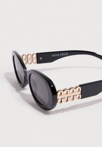 Μαύρα γυαλιά ηλίου με οβάλ φακούς, διακοσμημένα με χρυσή αλυσίδα στο πλάι. Γυαλιστερό φινίρισμα, κομψός σχεδιασμός, διακριτικά σκουρόχρωμοι φακοί.