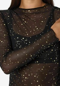 Prosojen črn top z zlatimi kovinskimi pikami, ki ima prilagojen kroj in dolge rokave. Mehka tekstura z zaobljenim vratnim izrezom in valovitimi manšetami.