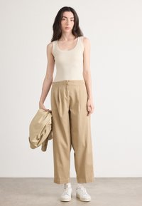 Beige tanktop, kaki wijde broek en witte sneakers. Het model houdt een bijpassende jas vast en staat op een lichtgrijze vloer.