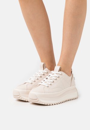 Nohy v béžových teniskách na platformě s tlustými texturovanými podrážkami a bílými tkaničkami na jednobarevném pozadí.