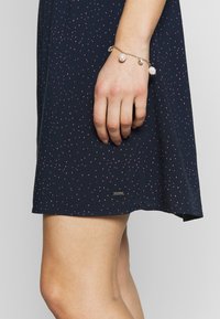 Abito navy con motivo a pois rosa; presenta una gonna corta e svasata. Bracciale in catena dorata con perle e charm su una mano vicino all'orlo.