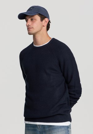Maglione lavorato a maglia blu navy con un motivo a coste testurizzato, collo rotondo e maniche lunghe, indossato sopra una camicia bianca; abbinato a un cappellino da baseball blu.