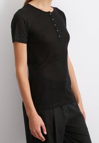 Top corto a maniche corte nero a costine con scollatura tonda e patta con quattro bottoni. Design aderente, superficie liscia e aspetto leggermente trasparente.