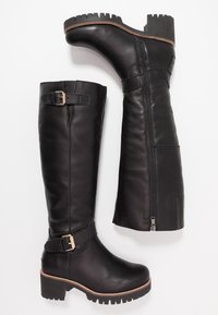 Botas de cuero negro hasta la rodilla con cremallera lateral, detalle de hebilla ajustable, suela gruesa y textura antideslizante en el tacón.