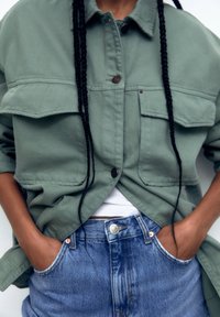 Light green jacket with two front pockets, worn over a white shirt. Paired with high-waisted blue denim shorts. Textured fabric visible.