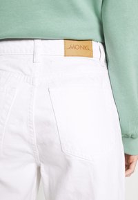 Valkoisetdenim-housut, joissa on tan-värinen nahkapaikka, jossa on teksti "MONKI" takana. Vyötärö ja taskut ovat näkyvissä, yhdistettynä mintunvihreään yläosaan.