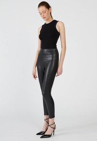 Czarna, bezrękawnikowa bluzka zestawiona z czarnymi, skórzanymi legginsami z wysokim stanem. Modelka nosi czarne, paskowe szpilki, prezentując elegancki, monochromatyczny look.