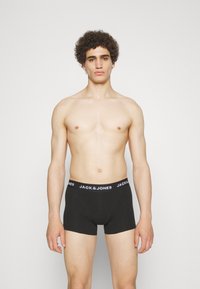 Schwarze Baumwollboxershorts mit einem weißen Logo-Bund. Glatte Textur, körpernahe Passform. Das Modell steht vor einem schlichten Hintergrund.