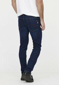 Jean en denim bleu foncé avec une coupe slim, présentant un motif de vague subtil sur les poches arrière et des surpiqûres contrastées sur les coutures.
